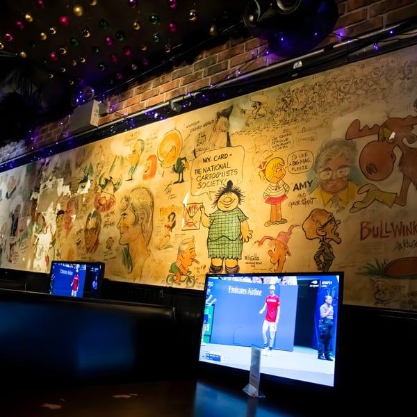 A sports bar wall covered in cartoon drawings, with two TVs showing a tennis match.