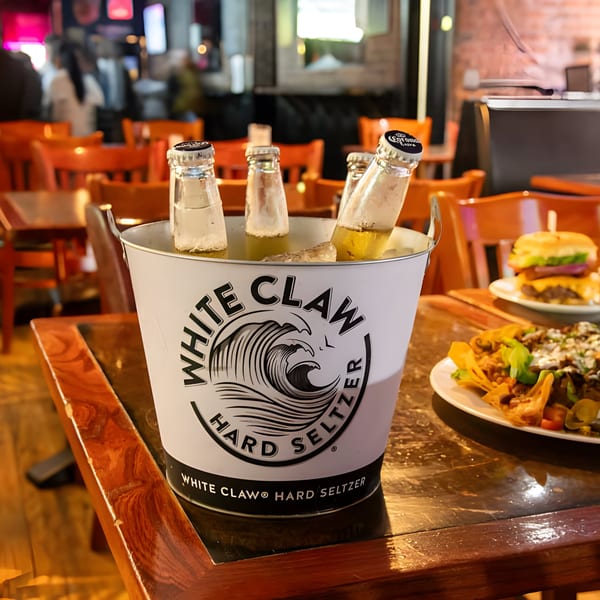 White Claw bucket with bottles and ice on a wooden table, with nachos and a burger in the background.