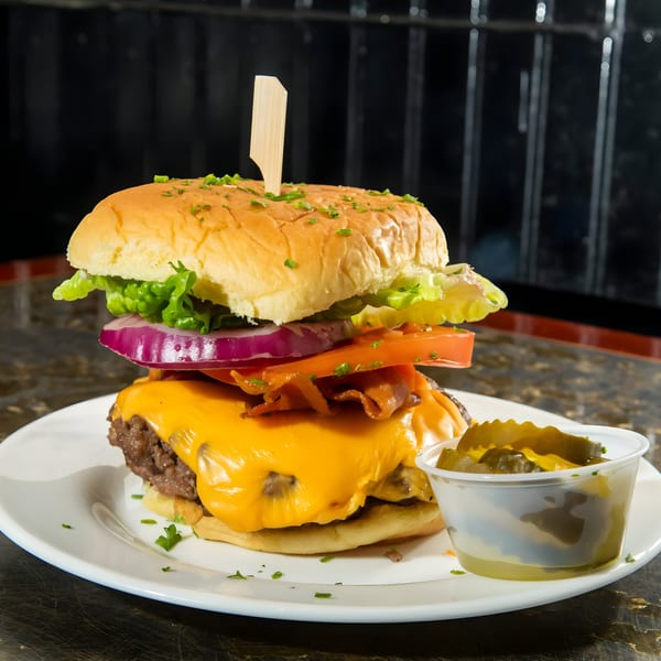 A tall cheeseburger with bacon, lettuce, tomato, and onion, served with a side of pickles.