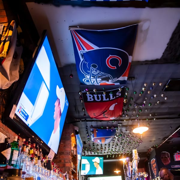 Sports bar interior with multiple TVs showing a game, liquor bottles, and team flags hanging from the ceiling.
