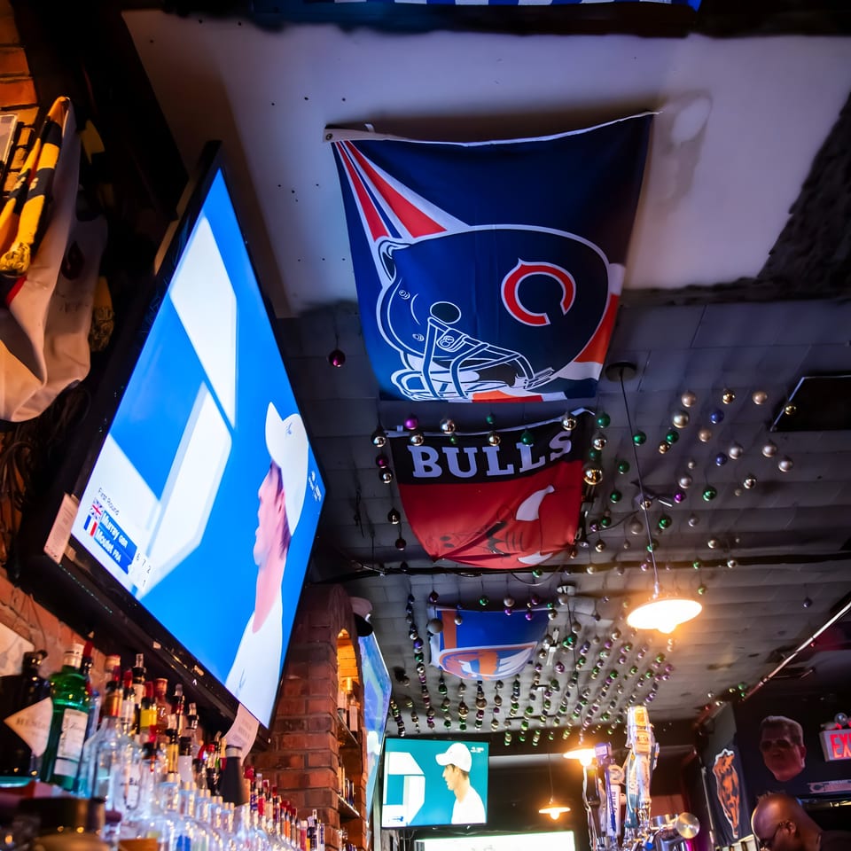 Sports bar interior with multiple TVs showing a game, liquor bottles, and team flags hanging from the ceiling.