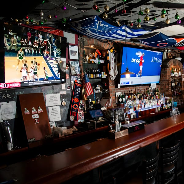 A sports bar interior with two large TVs showing games, a fully stocked bar, and Chicago Bears flags.