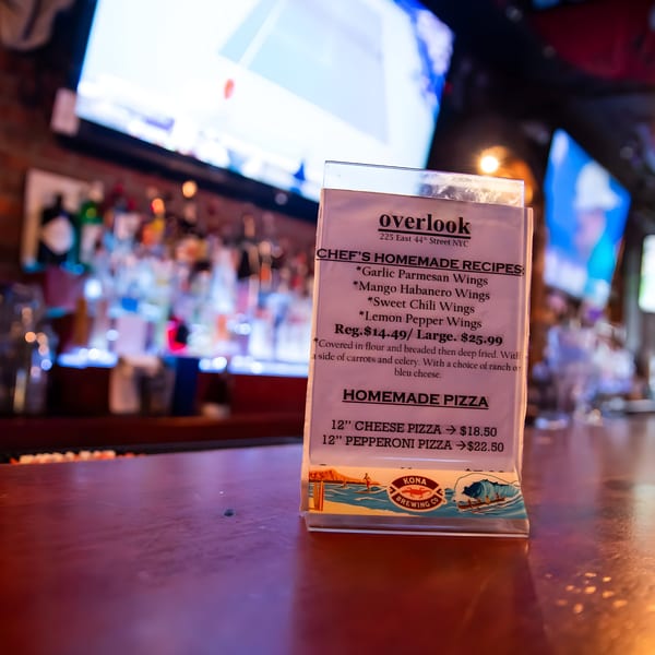 A menu for Overlook sports bar, featuring wings and pizza, on a wooden bar with blurred bottles and a TV in the background.