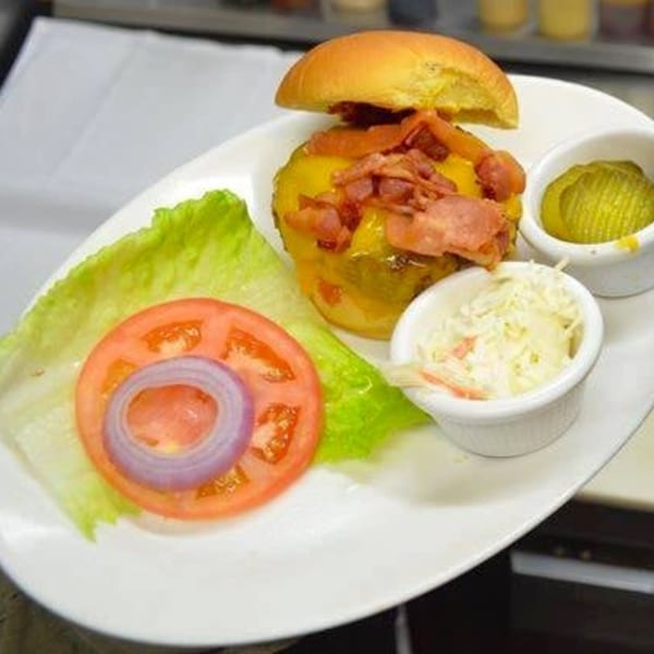A bacon cheeseburger with lettuce, tomato, onion, coleslaw, and pickles on a white plate.